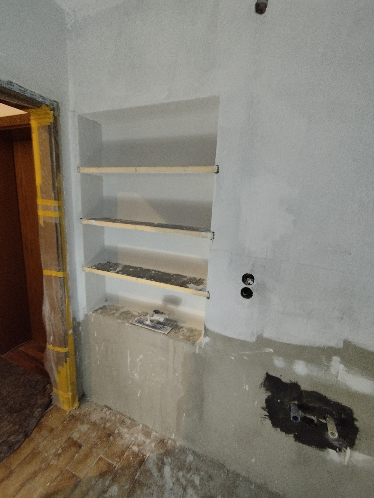 Narożnik ściany z wnęką na półki w trakcie szpachlowania. Widoczne gniazdka elektryczne i rury. Podłoga częściowo zakryta folią. Prace wykończeniowe w szarym odcieniu.