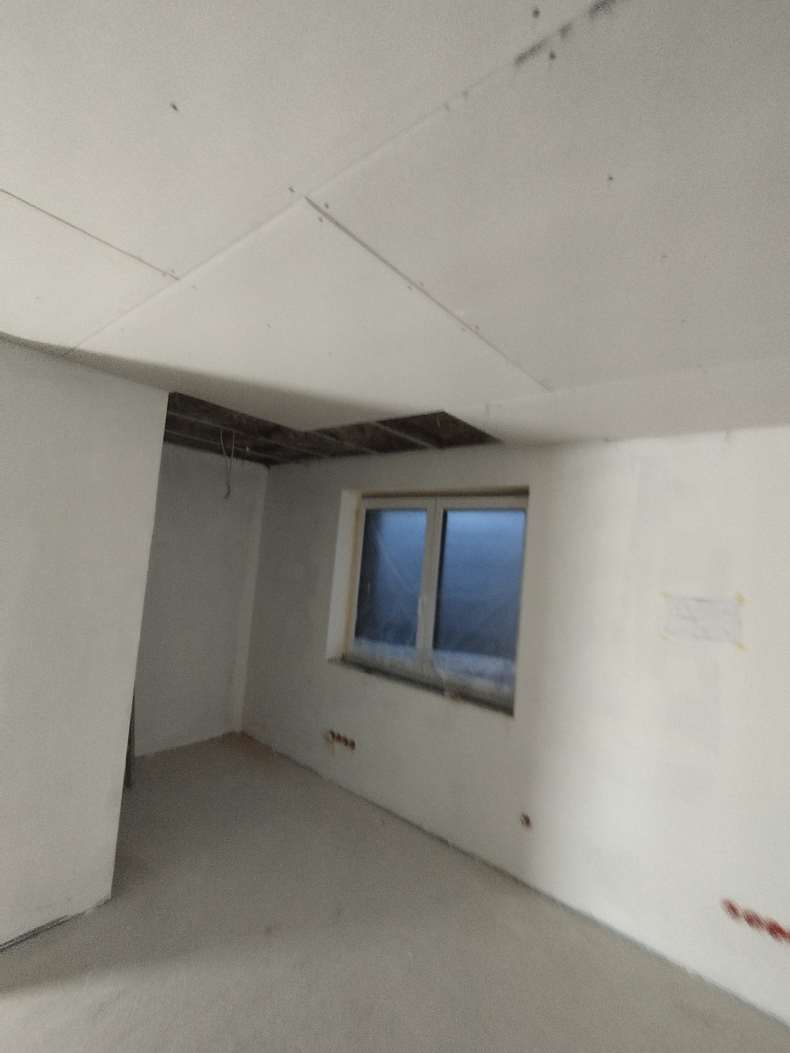 Wnętrze w trakcie remontu: widoczne ściany, okno, sufit z częściowo zamontowanymi płytami gipsowymi i odsłoniętą konstrukcją. Na ścianie widoczne ślady po instalacjach.