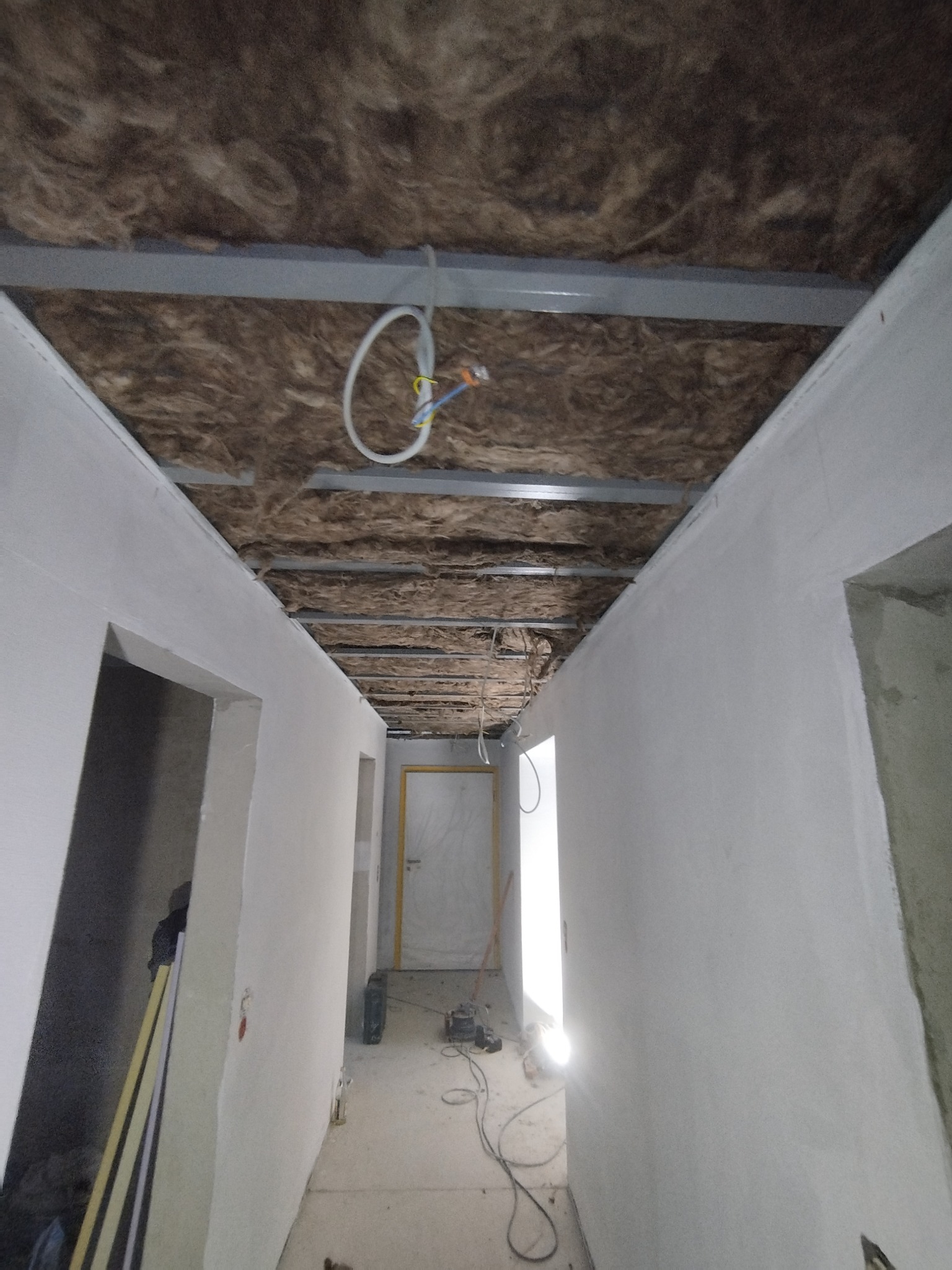 Korytarz w trakcie prac wykończeniowych: ocieplony sufit z widoczną wełną mineralną i metalową konstrukcją, ściany przygotowane do malowania, widoczne kable elektryczne.