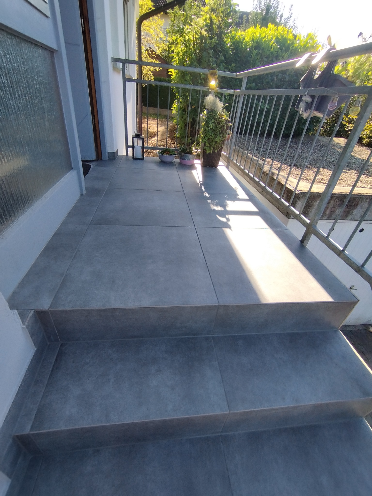 Wykończony taras z szarymi płytkami, widoczne schody i metalowa balustrada. Słoneczny dzień, rośliny w tle. Minimalistyczny design.