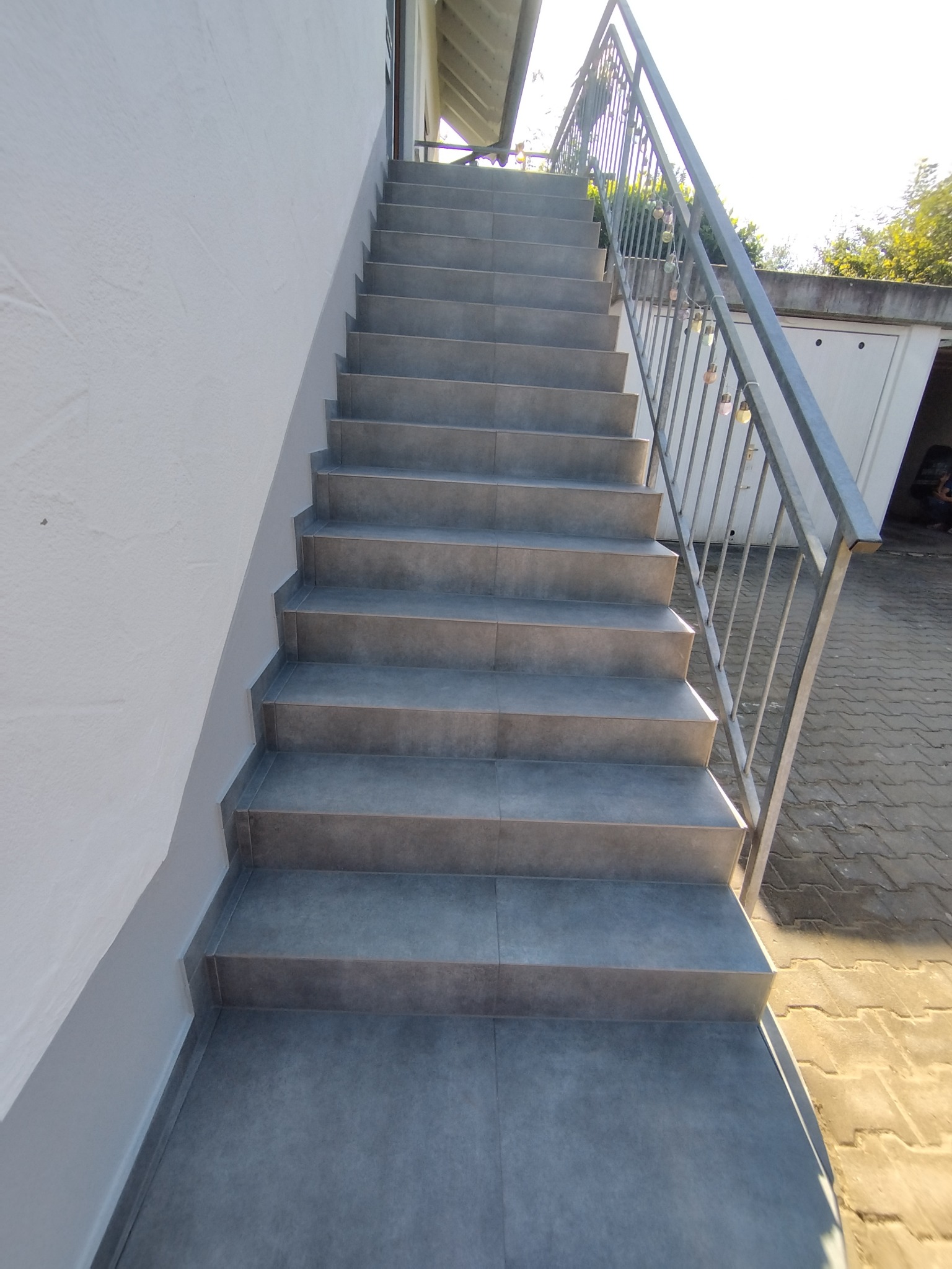 Zewnętrzne schody wyłożone szarymi płytkami, prowadzące do góry, z metalową barierką po prawej stronie. Widok z dołu, jasne oświetlenie.