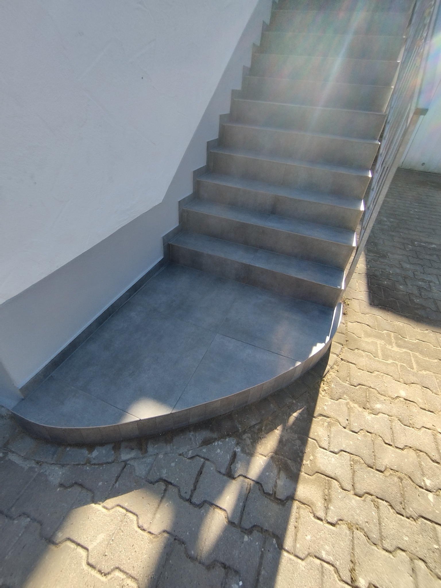 Wykończone płytkami schody zewnętrzne z zaokrąglonym podestem, wpadające w kadr promienie słoneczne, szara kolorystyka, kostka brukowa na dole kadru.