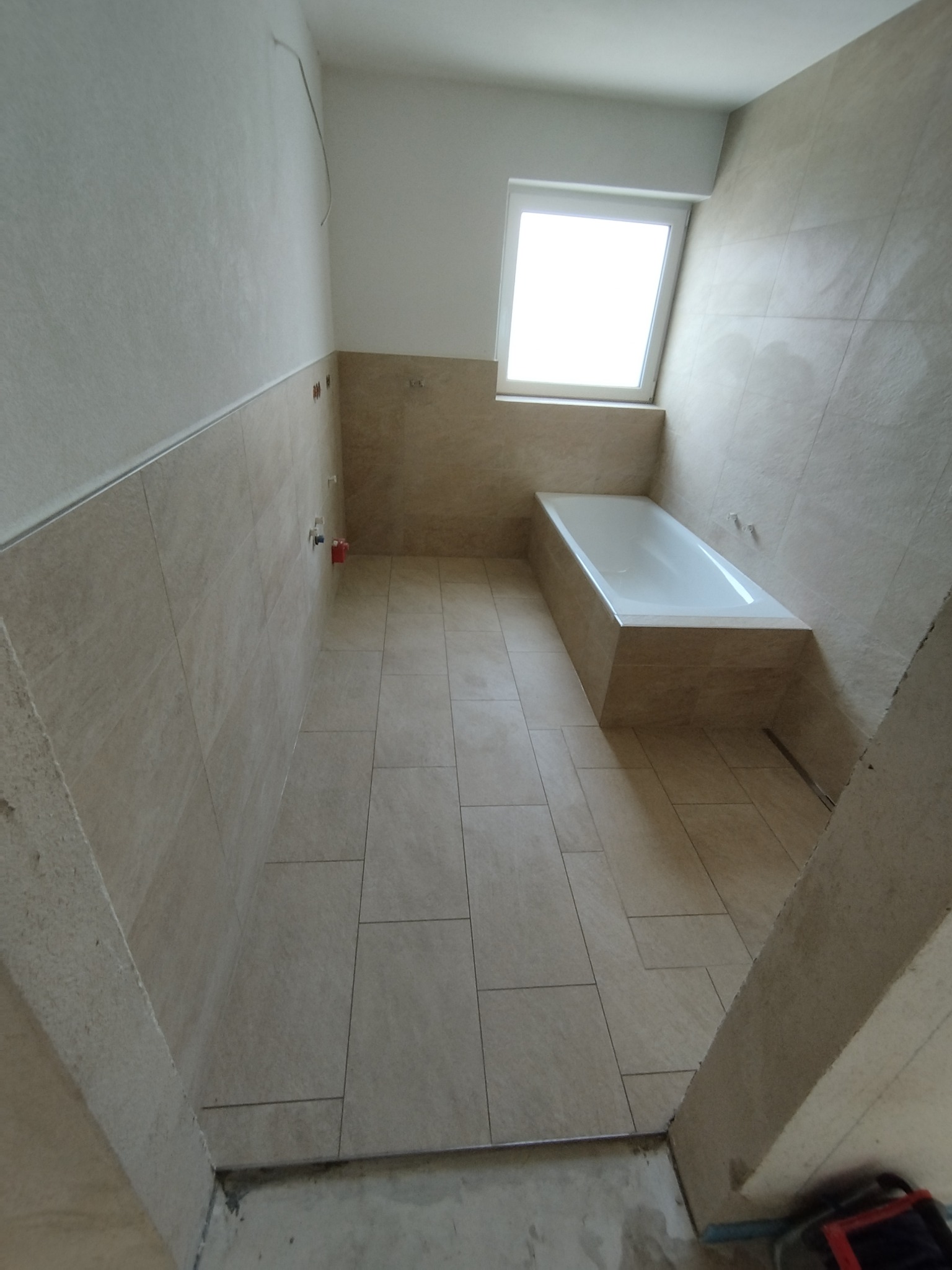 Jasna łazienka po remoncie: beżowe płytki na ścianach i podłodze, biała wanna pod oknem. Widoczne niedokończone detale przy podłodze i ścianach.