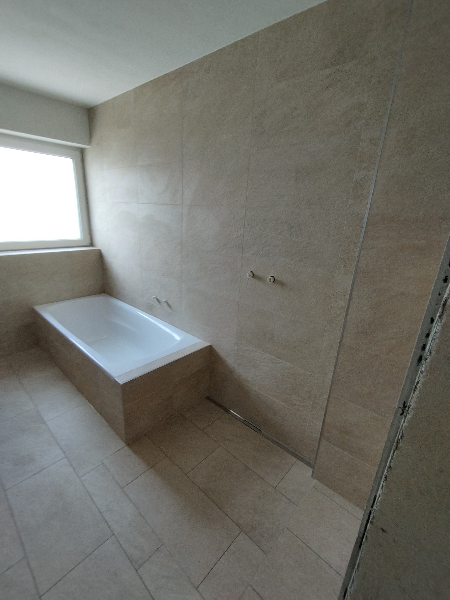 Jasna łazienka z beżowymi płytkami, białą wanną i oknem. Widoczne przyłącza do baterii. Minimalistyczny design, świeżo po remoncie.