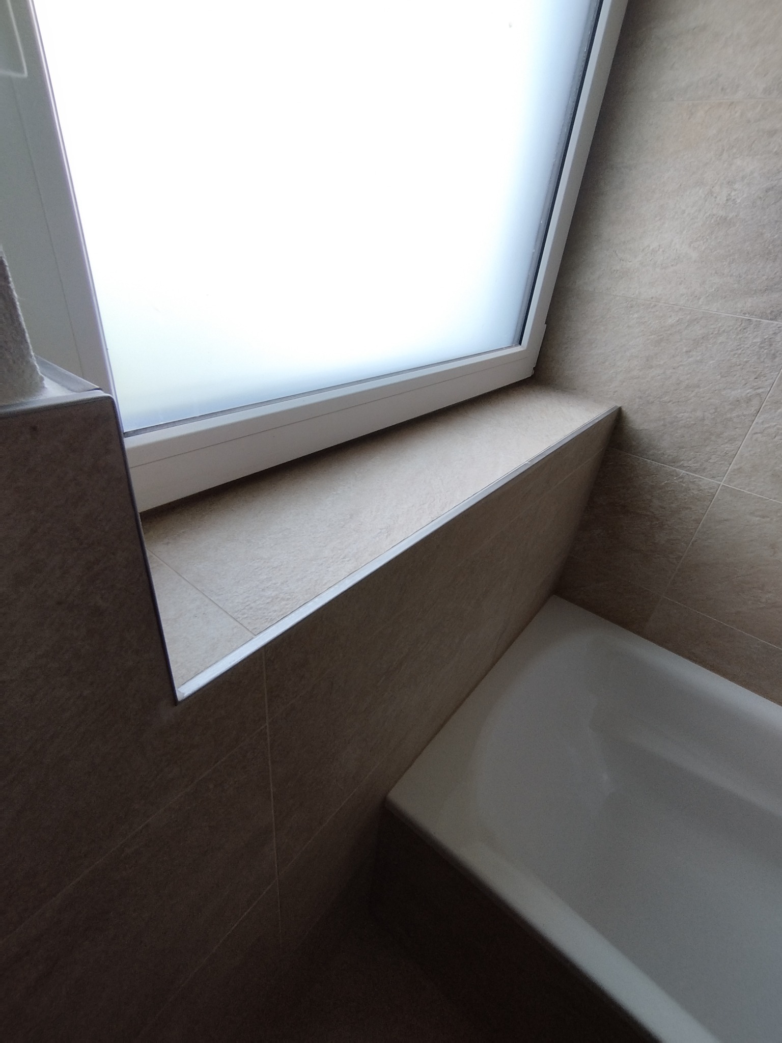 Beżowe płytki w łazience z oknem i wanną. Widoczny fragment wanny, parapet z płytek i okno z mleczną szybą. Minimalistyczny design.