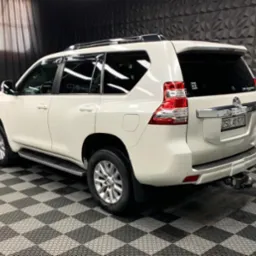 Biały samochód Toyota Land Cruiser Prado po detailingu, widok z tyłu i boku w studiu z czarno-białą podłogą w szachownicę i ciemną ścianą z geometrycznym wzorem, widoczne naklejki na tylnej szybie.