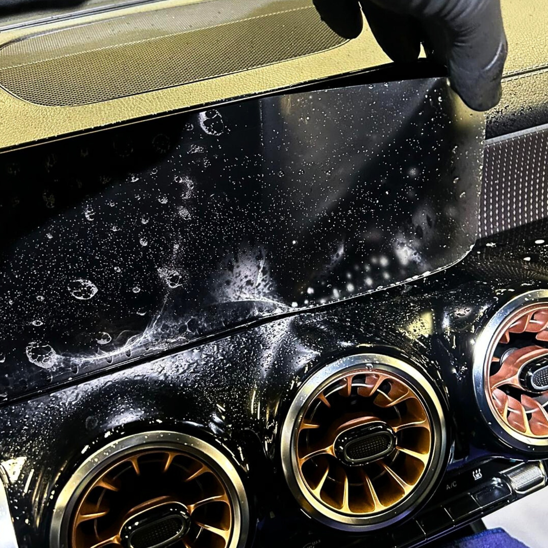 Detailing wnętrza samochodu: czyszczenie mokrej, czarnej deski rozdzielczej z okrągłymi, złotymi kratkami wentylacyjnymi przez osobę w czarnej rękawicy.