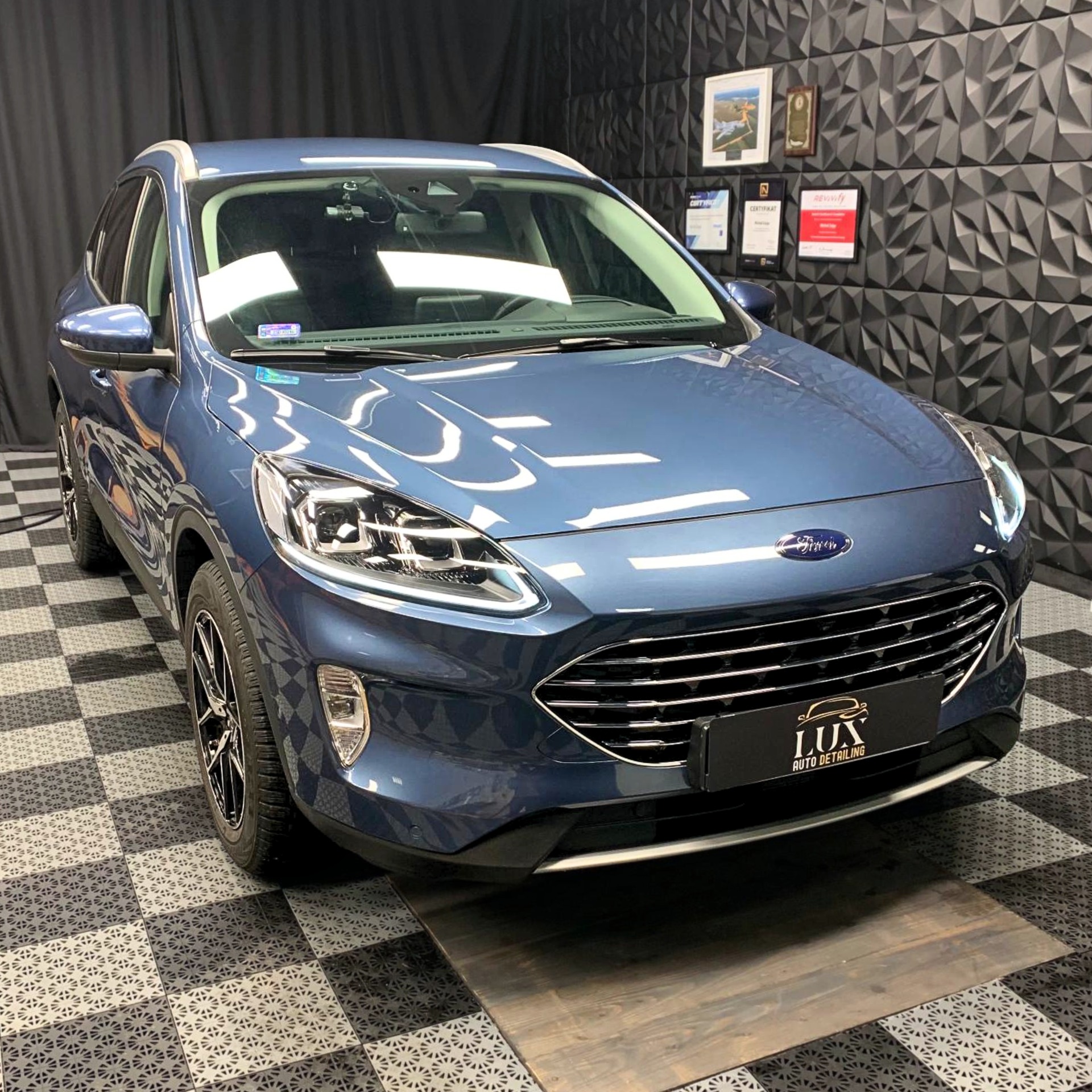 Błękitny Ford Kuga po zabiegach auto detailingu, prezentowany w studiu z czarno-białą szachownicą na podłodze i trójwymiarową ścianą w tle, z widocznym logo 'LUX Auto Detailing'.