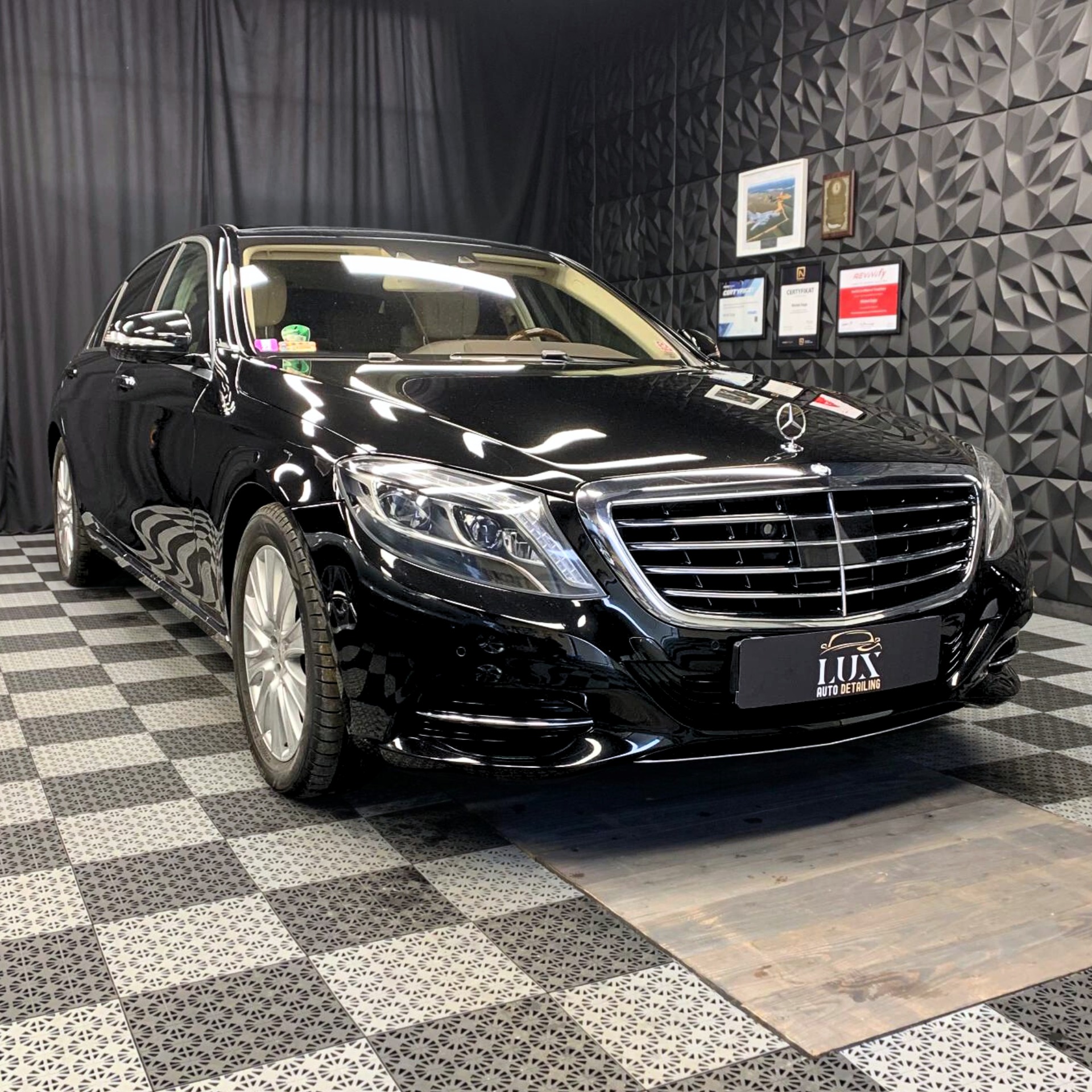 Błyszczący, czarny Mercedes-Benz S-Class po zabiegach auto detailingu, zaparkowany w studiu z dekoracyjną, trójwymiarową ścianą i szachownicą na podłodze. Na przednim zderzaku widoczna tabliczka...
