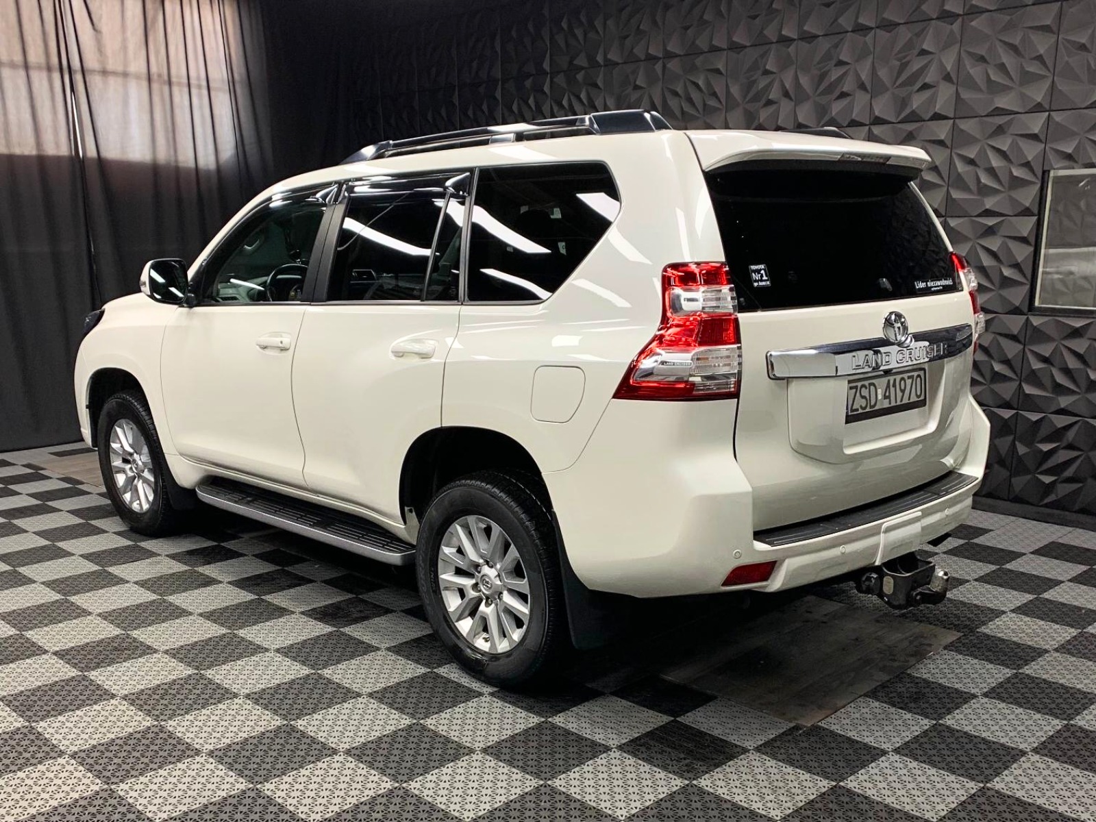 Biały samochód Toyota Land Cruiser Prado po detailingu, widok z tyłu i boku w studiu z czarno-białą podłogą w szachownicę i ciemną ścianą z geometrycznym wzorem, widoczne naklejki na tylnej szybie.