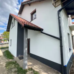 Wykończona elewacja domu jednorodzinnego z białym tynkiem strukturalnym, grafitowymi akcentami na cokole i ścianie szczytowej, oraz czerwoną dachówką ceramiczną, widoczne rynny i fragment chodnika...