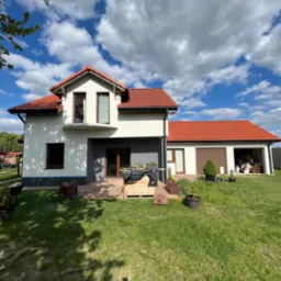 Dwukondygnacyjny dom z czerwoną dachówką i świeżo pomalowaną elewacją w odcieniach bieli i szarości, widok z trawnika w słoneczny dzień z dekoracyjnymi roślinami w donicach.