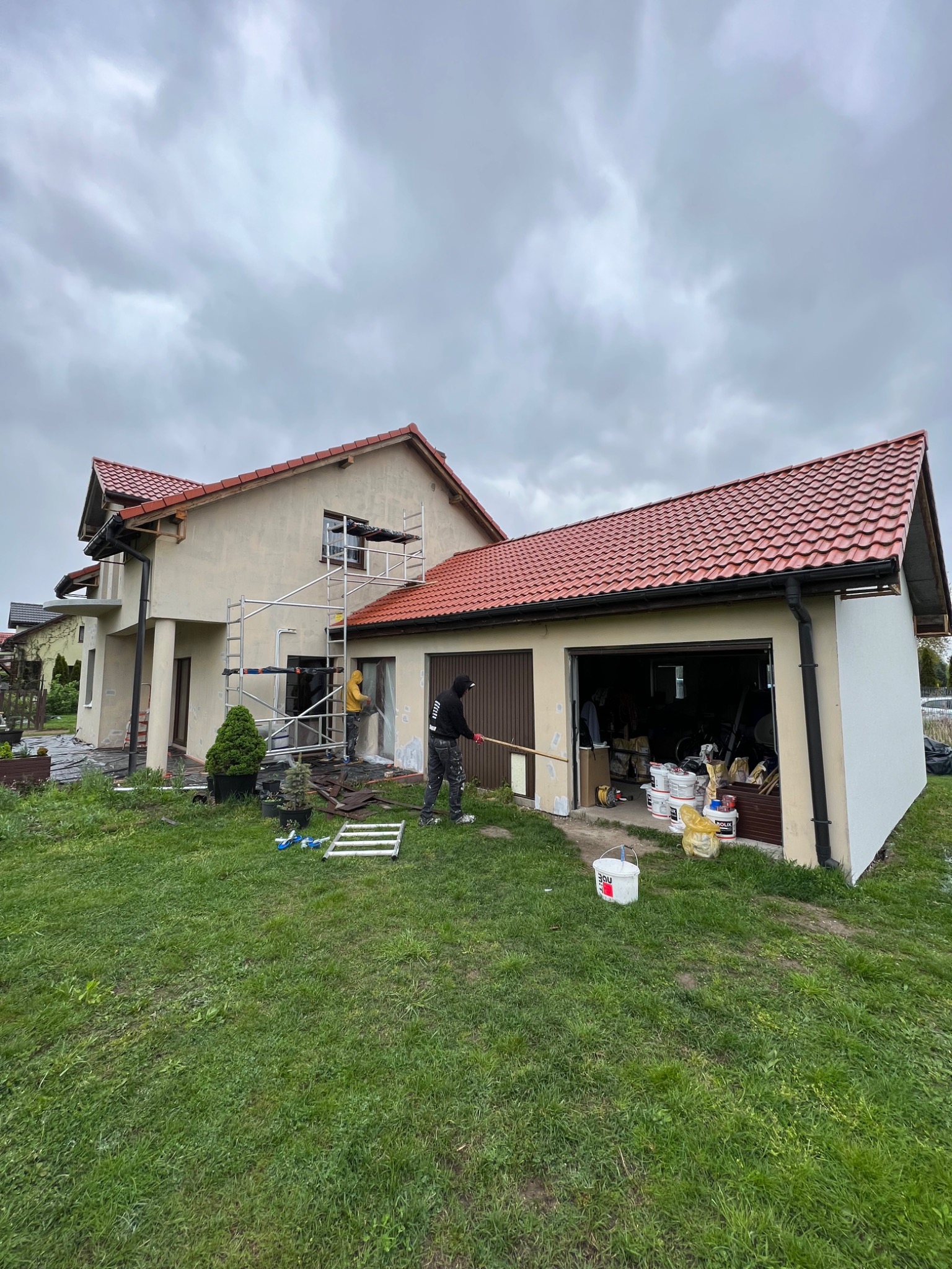 Dwóch pracowników na rusztowaniu i z wałkiem malarskim wykonuje prace na elewacji domu jednorodzinnego z czerwonym dachem w pochmurny dzień; widoczne liczne puszki z farbą i narzędzia.