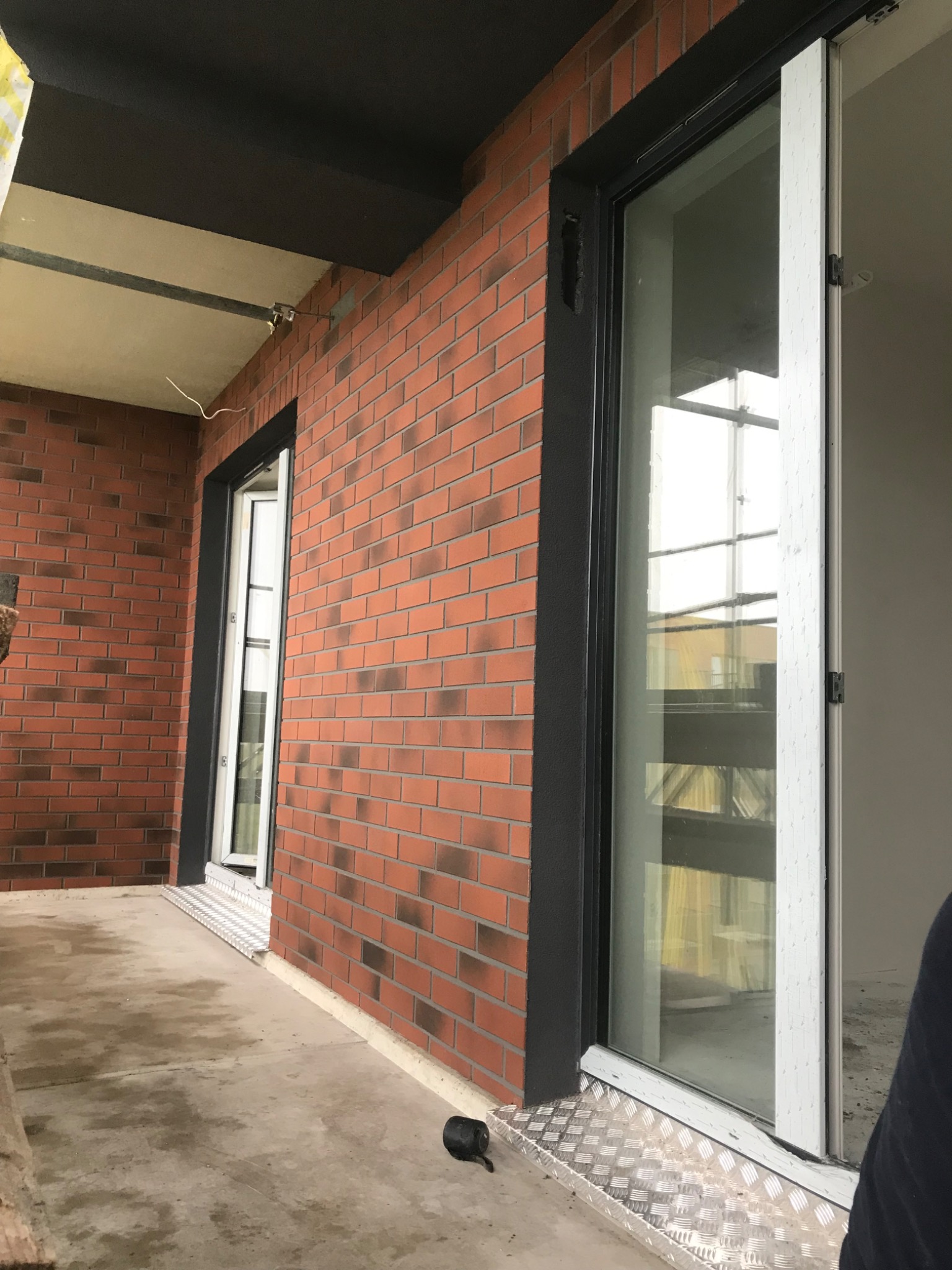 Elewacja budynku wykończona czerwoną cegłą klinkierową z ciemniejszymi przebarwieniami, widoczne okno z czarną ramą i białą ramą okienną, aluminiowy próg, surowy betonowy balkon.