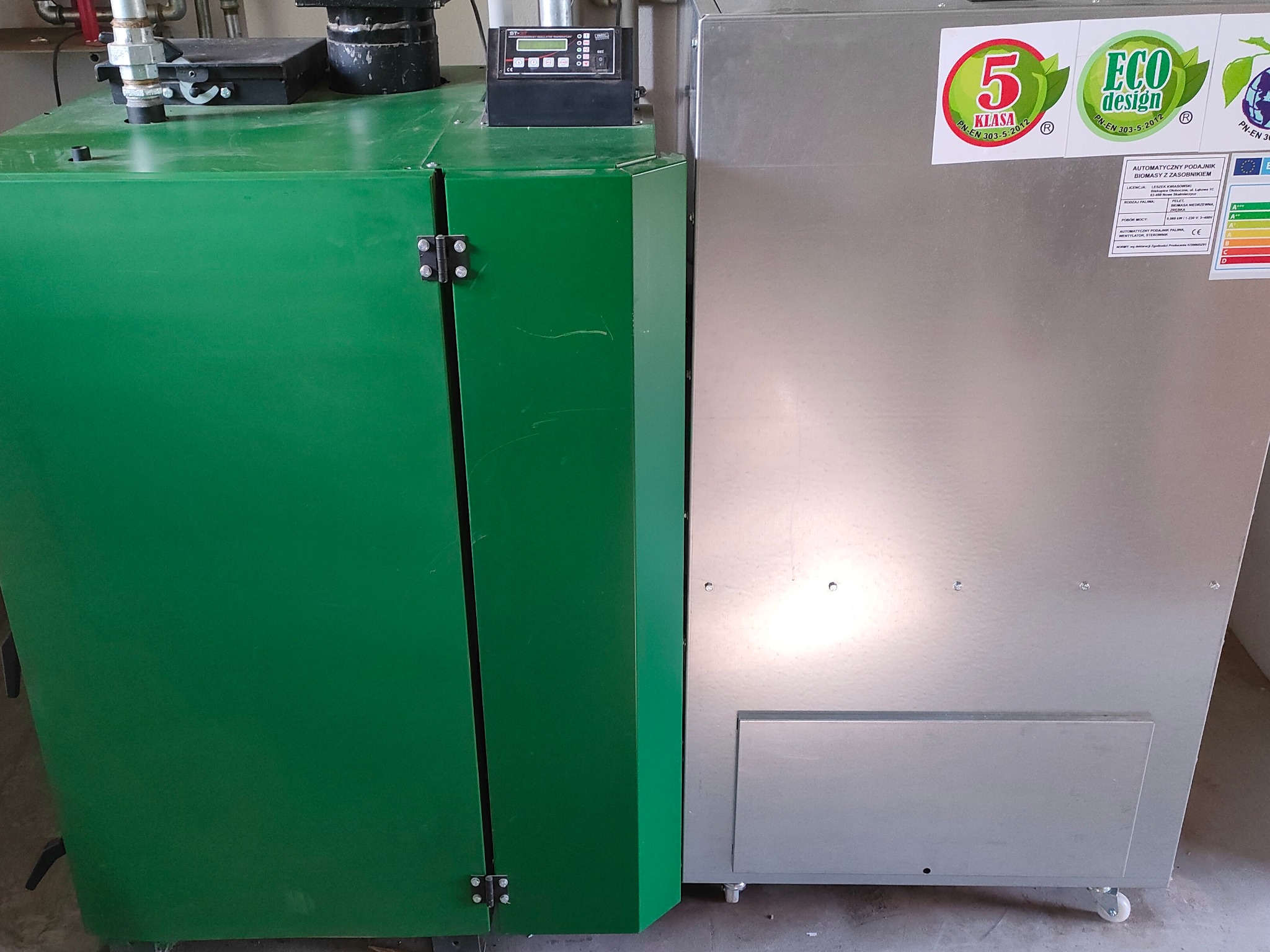 Nowoczesny kocioł na paliwo stałe z automatycznym podajnikiem, widoczne oznaczenia klasy energetycznej i ekologicznego designu, obudowa w kolorze zielonym i srebrnym.
