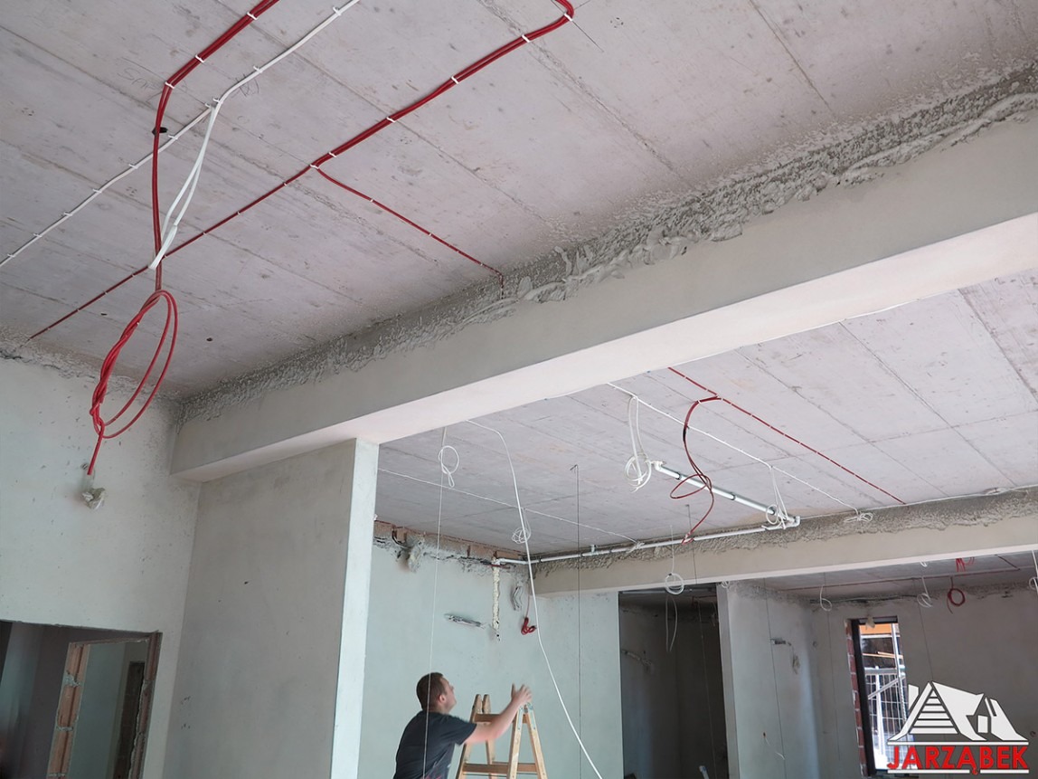Instalacja elektryczna w surowym wnętrzu: czerwone i białe przewody prowadzone po betonowym suficie, elektryk na drabinie w tle.