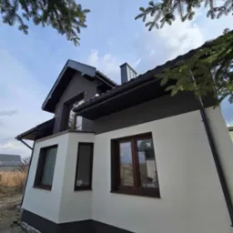 Dwukondygnacyjny dom z jasną elewacją, ciemnym dachem i brązowymi oknami. Ujęcie z dołu, fragment drzewa w prawym górnym rogu. Nowoczesny styl.