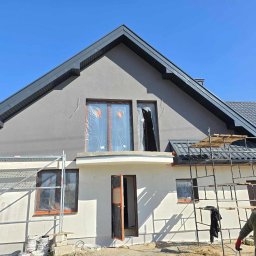 DaRo Bud Dariusz Kucab - Dom jednorodzinny w trakcie malowania elewacji na szaro, z rusztowaniem i zabezpieczonymi oknami folią. Widoczny fragment osoby w prawym dolnym rogu.