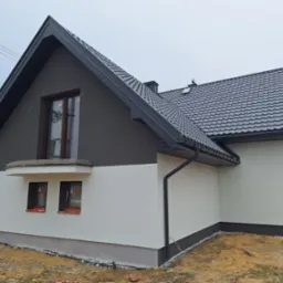 Dwukolorowa elewacja domu jednorodzinnego z szarą górą i białą dolną częścią. Widoczne okna z brązowymi ramami i fragment dachu z czarną dachówką.