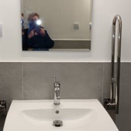 DaRo Bud Dariusz Kucab - Biała umywalka z chromowaną baterią i uchwytem dla niepełnosprawnych na tle szarej ściany i lustra z odbiciem osoby robiącej zdjęcie w łazience.