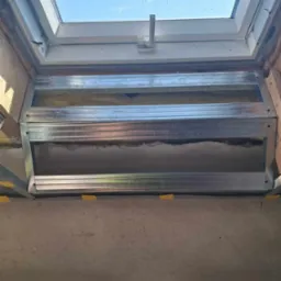 Montaż stelaża pod sufit napinany wokół okna dachowego, widoczne profile metalowe i częściowo wypełniona przestrzeń izolacją. Ujęcie z dołu, w tle błękitne niebo.