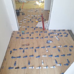 DaRo Bud Dariusz Kucab - Układanie paneli podłogowych z użyciem klipsów poziomujących. Widoczny fragment podłogi z narzędziami i materiałami budowlanymi w tle. Jasne wnętrze w trakcie prac wykończeniowych.