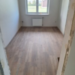 DaRo Bud Dariusz Kucab - Pokój po remoncie z nową, brązową podłogą z paneli, widok zza futryny. Ściany pomalowane na biało, okno z widokiem na zieleń. Minimalistyczny grzejnik pod oknem.