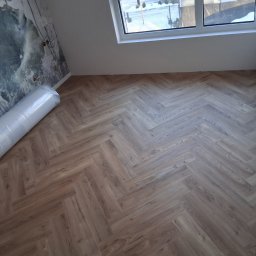 DaRo Bud Dariusz Kucab - Ukończone układanie paneli podłogowych w jodełkę w pokoju na poddaszu z oknem dachowym. Rolka paneli przy ścianie z tapetą w kwiaty.