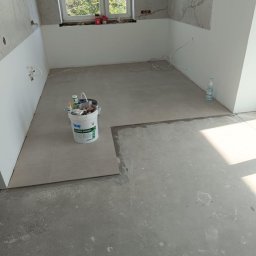 DaRo Bud Dariusz Kucab - Wnętrze kuchni w trakcie remontu: częściowo ułożone płytki podłogowe, wiadro z zaprawą Knauf Super Finish, okno z widokiem na zieleń. Prace wykończeniowe w toku.