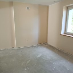 DaRo Bud Dariusz Kucab - Jasne, świeżo pomalowane wnętrze pokoju z oknem i surową podłogą. Widoczny nawiewnik na ścianie i listwa przypodłogowa. Minimalistyczny styl, neutralna kolorystyka.