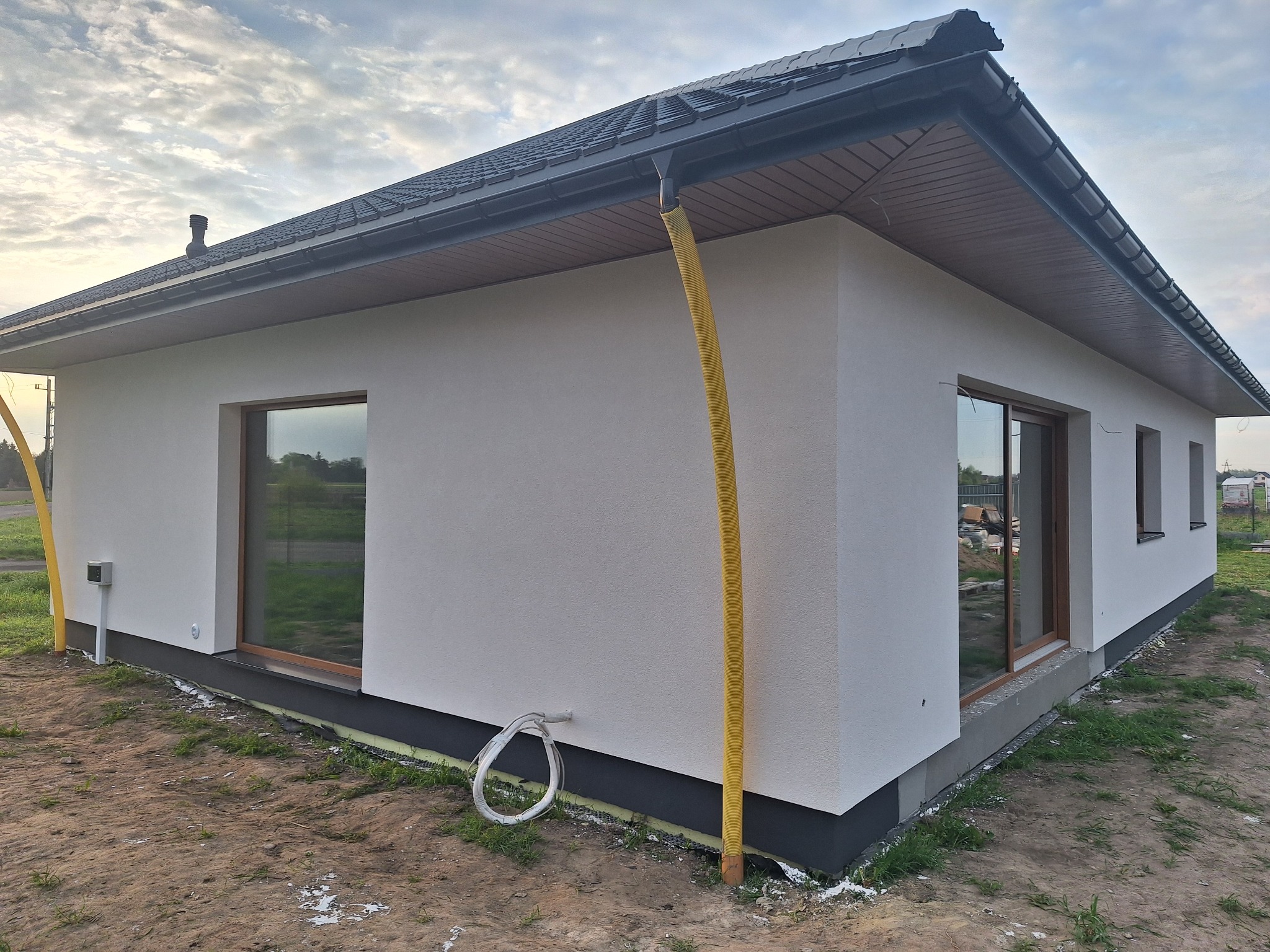 Nowoczesny dom jednorodzinny z ocieploną białą elewacją, grafitową podbitką i rynnami. Żółty przewód wentylacyjny na ścianie. Okna z drewnianą stolarką.