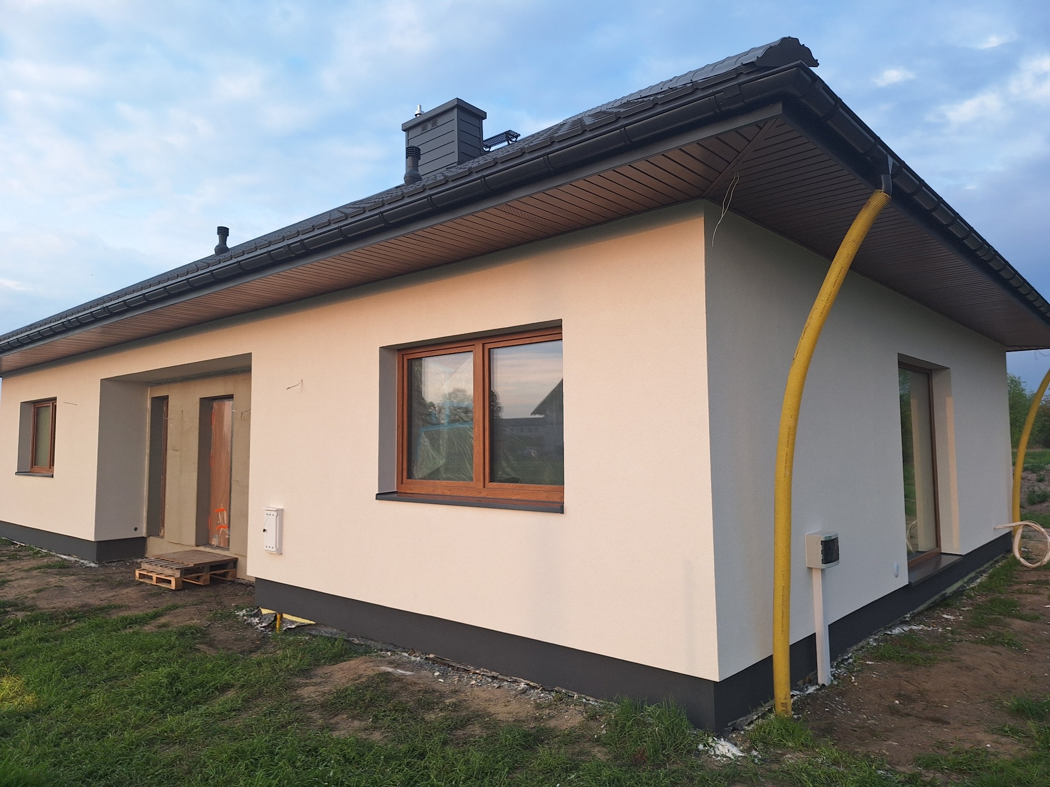 Otynkowany dom jednorodzinny z brązowymi ramami okiennymi, szarą podbitką i cokołem. Żółty wąż wentylacyjny elewacji. Widok z zewnątrz na świeżo otynkowaną elewację.
