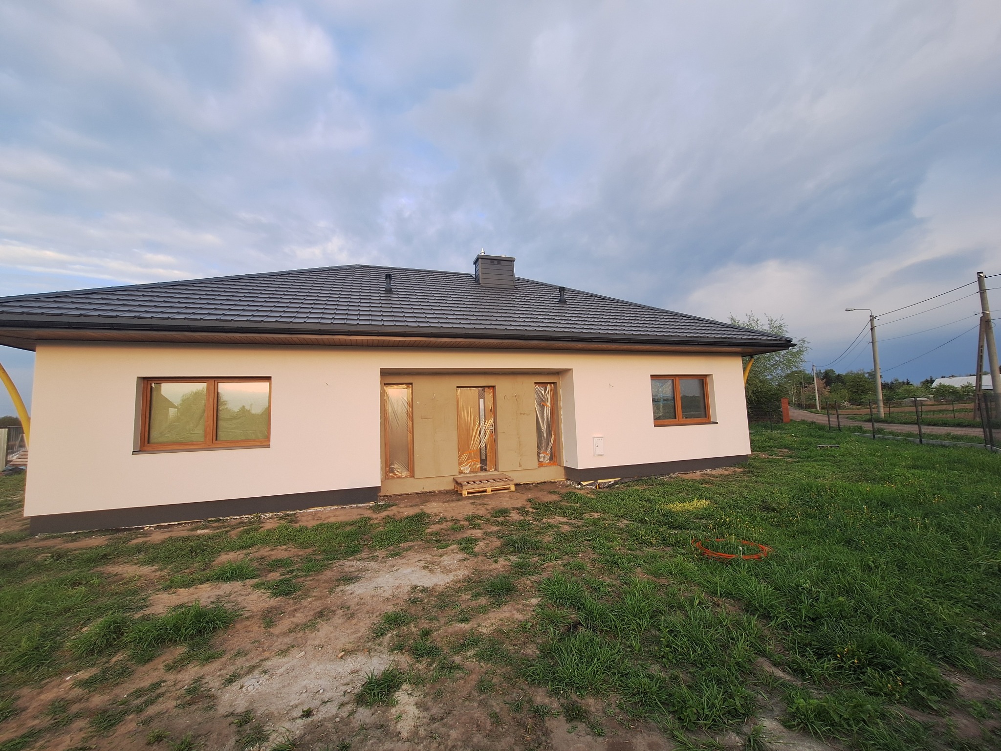 Biały dom z ciemnym dachem w trakcie wykończenia elewacji. Okna z drewnianą ramą. Wejście zabezpieczone. Widok na niebo z chmurami.