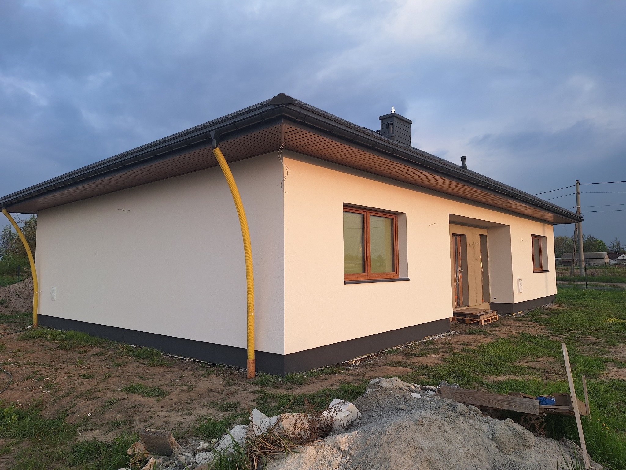 Nowo otynkowany, parterowy dom z brązowymi oknami, ciemną podmurówką i żółtymi rurami spustowymi w promieniach zachodzącego słońca.