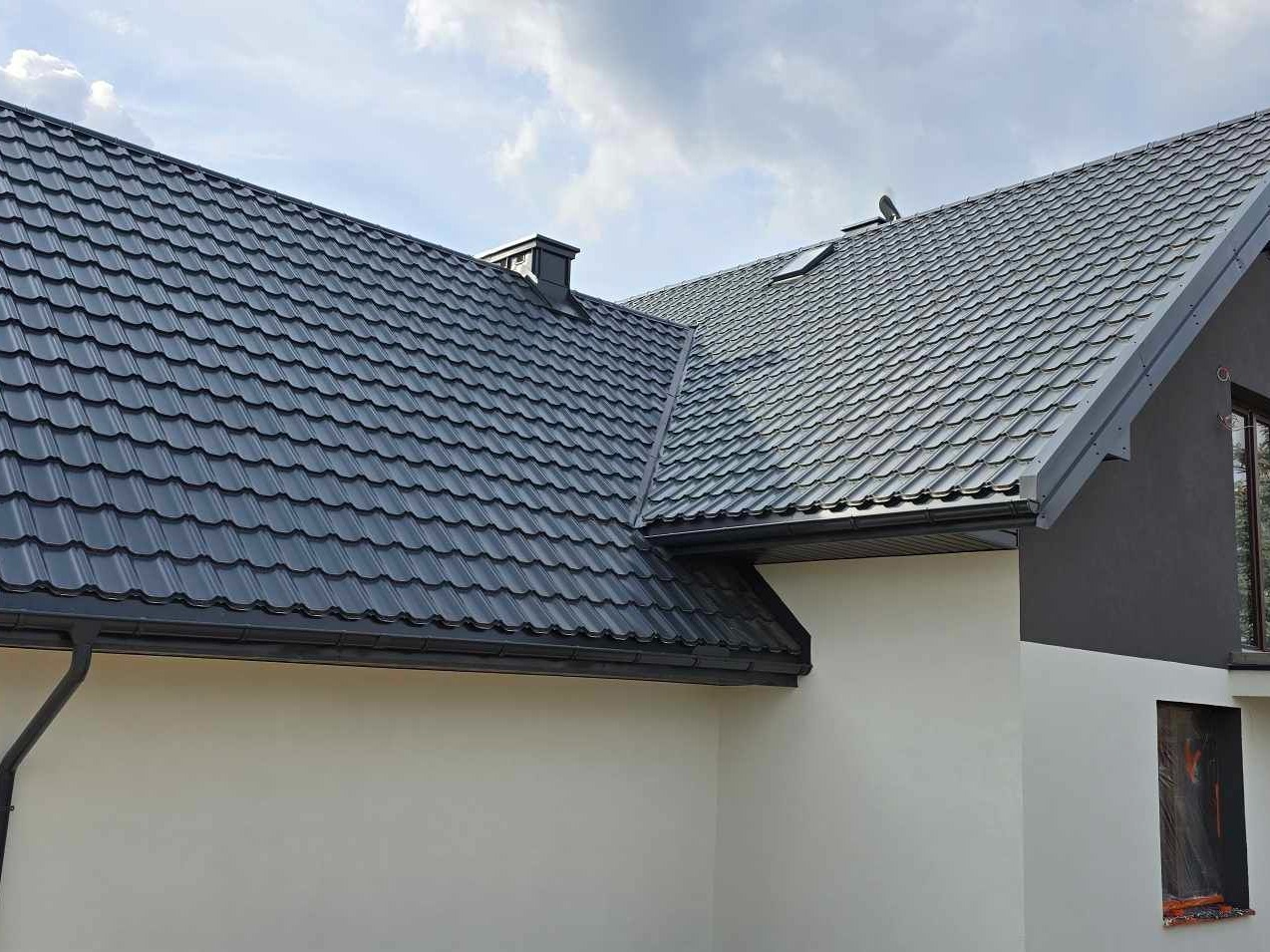 Szara blachodachówka na domu jednorodzinnym w Podkampinos. Widoczny fragment elewacji w dwóch odcieniach szarości i okno zabezpieczone folią.
