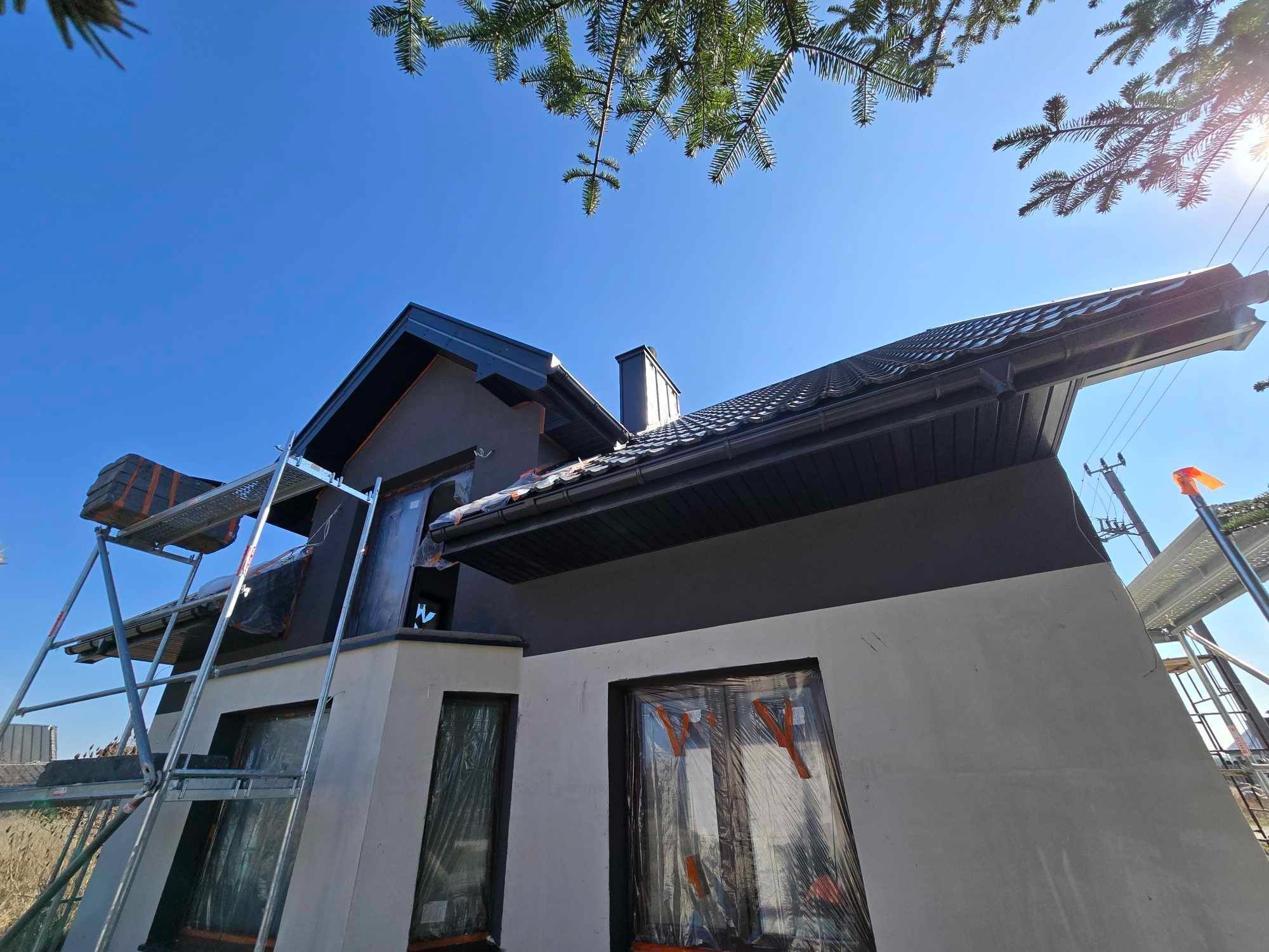 Dom w trakcie ocieplania elewacji z użyciem rusztowania. Okna zabezpieczone folią. Widok z dołu na dwukolorową elewację i błękitne niebo.