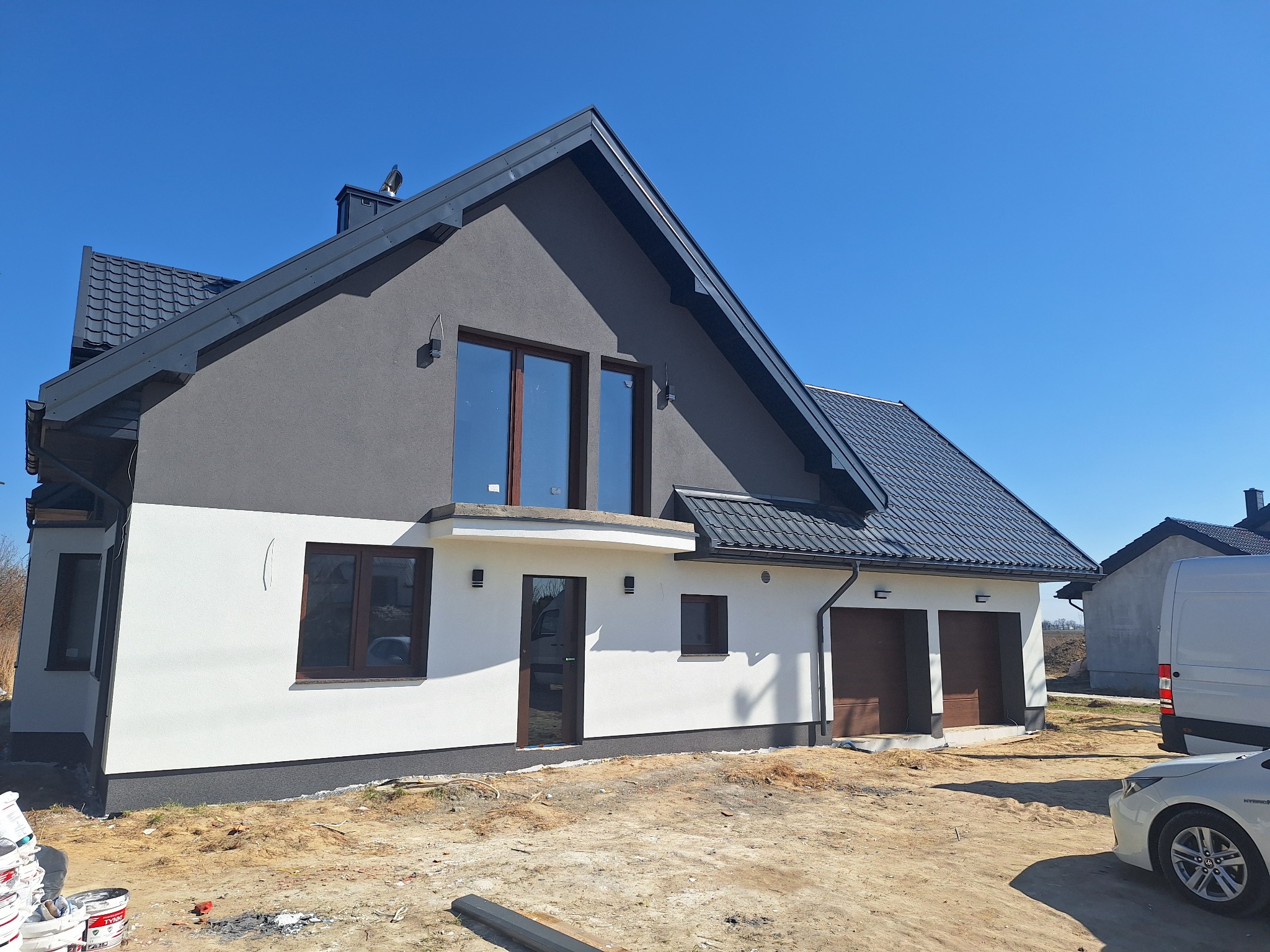 Dwukondygnacyjny dom z szarą elewacją na górze i białą na dole, z brązowymi oknami i drzwiami garażowymi, na tle błękitnego nieba w słoneczny dzień.