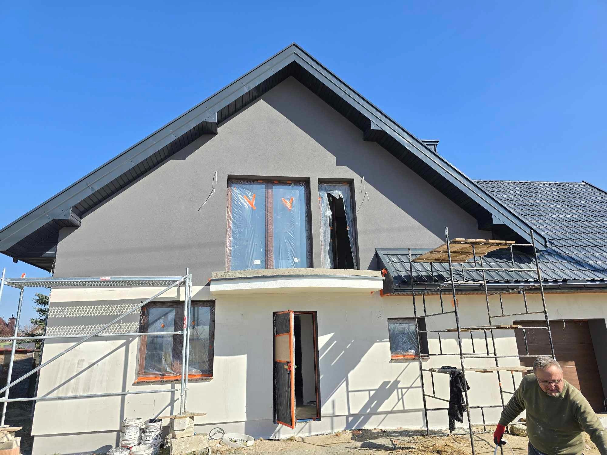 Dom jednorodzinny w trakcie malowania elewacji na szaro, z rusztowaniem i zabezpieczonymi oknami folią. Widoczny fragment osoby w prawym dolnym rogu.