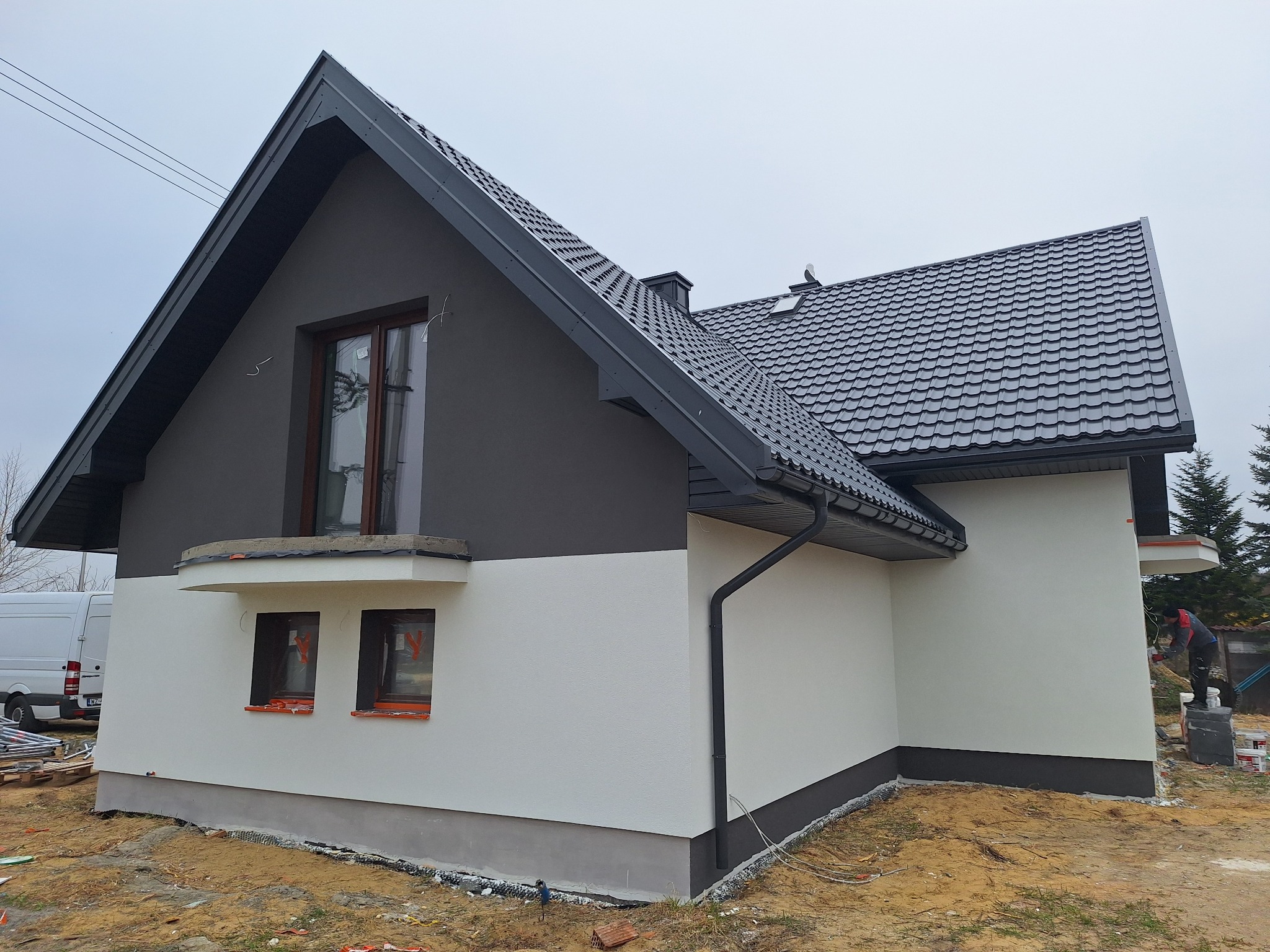 Dwukolorowa elewacja domu jednorodzinnego z szarą górą i białą dolną częścią. Widoczne okna z brązowymi ramami i fragment dachu z czarną dachówką.