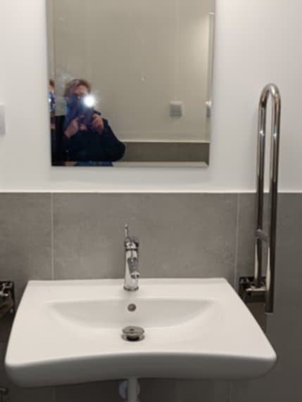 Biała umywalka z chromowaną baterią i uchwytem dla niepełnosprawnych na tle szarej ściany i lustra z odbiciem osoby robiącej zdjęcie w łazience.