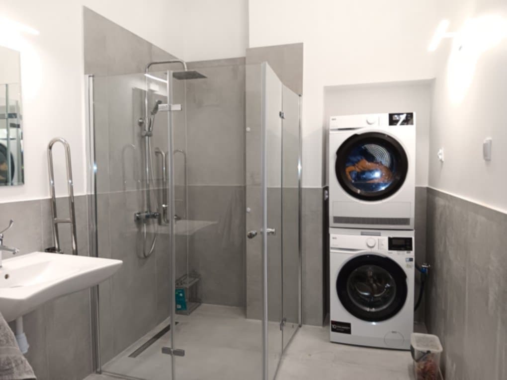 Nowoczesna łazienka z szarą kabiną prysznicową typu walk-in, umywalką i pralko-suszarką w słupku. Minimalistyczny design z szarymi płytkami i białymi ścianami.