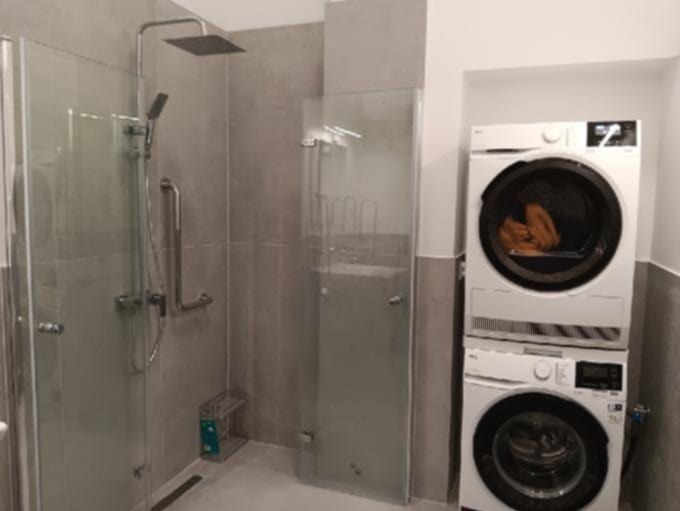 Nowoczesna łazienka z przeszklonym prysznicem typu walk-in, szarymi płytkami i pralko-suszarką w zabudowie pionowej. Minimalistyczny design, funkcjonalne rozwiązania.