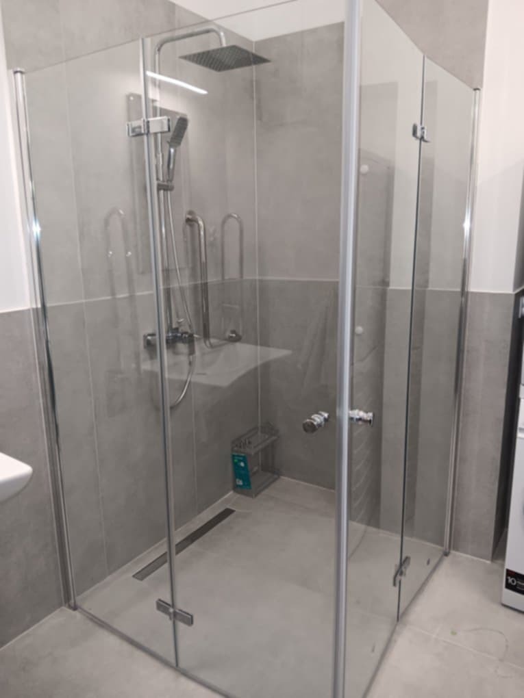 Szklana kabina prysznicowa z deszczownicą i uchwytami, wykończona szarą płytką. Widoczny odpływ liniowy, minimalistyczny design. Nowoczesna aranżacja łazienki.