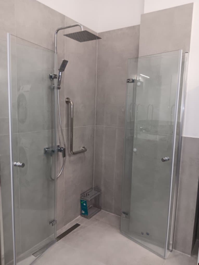 Nowoczesna kabina prysznicowa z deszczownicą i szklanymi drzwiami, wykończona szarą płytką. Widoczna armatura i uchwyty dla osób z niepełnosprawnościami.