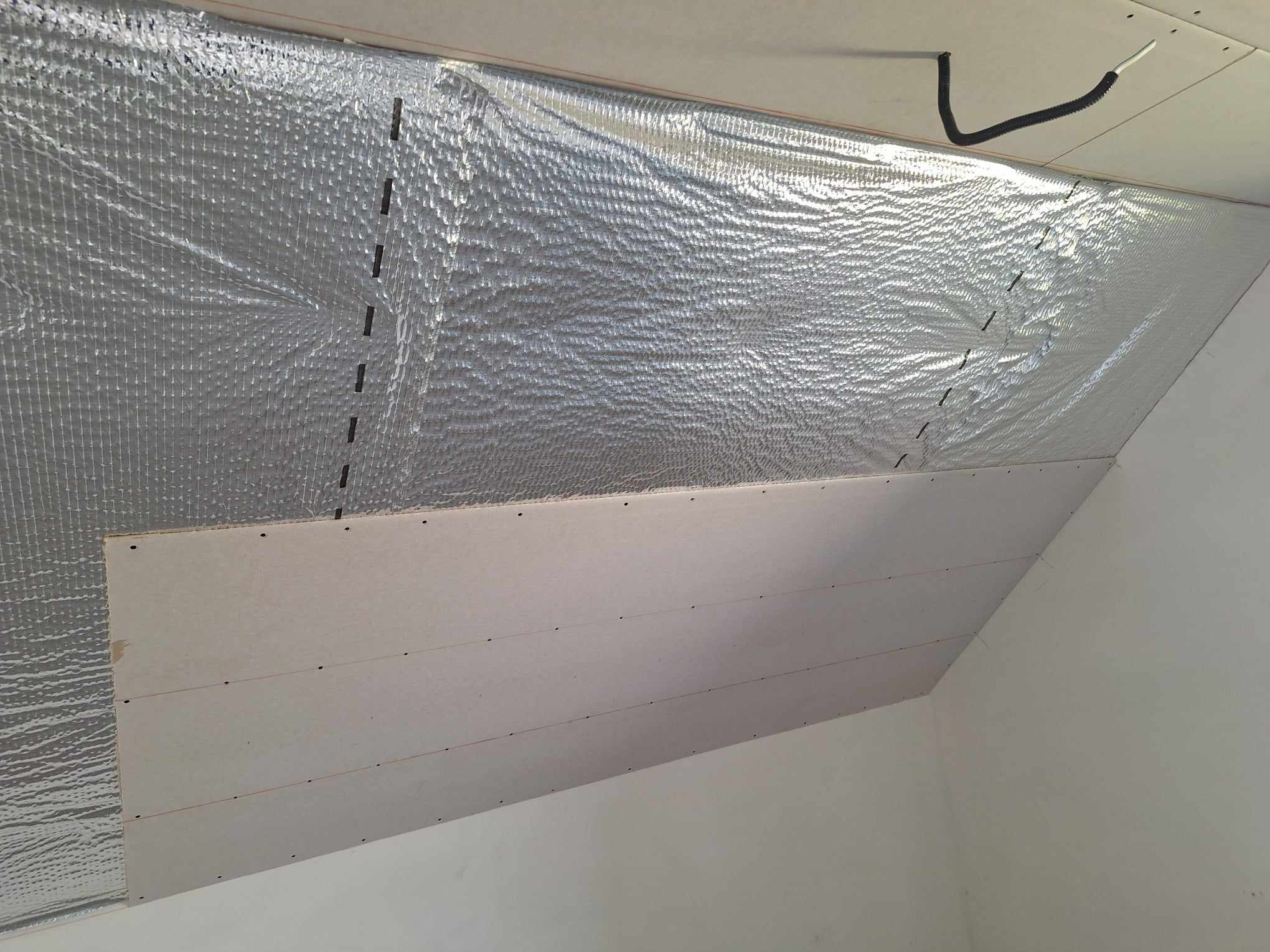 Izolacja poddasza z folią aluminiową i płytą gipsową, widok z dołu. Czarny kabel przy suficie. Etap prac wykończeniowych.