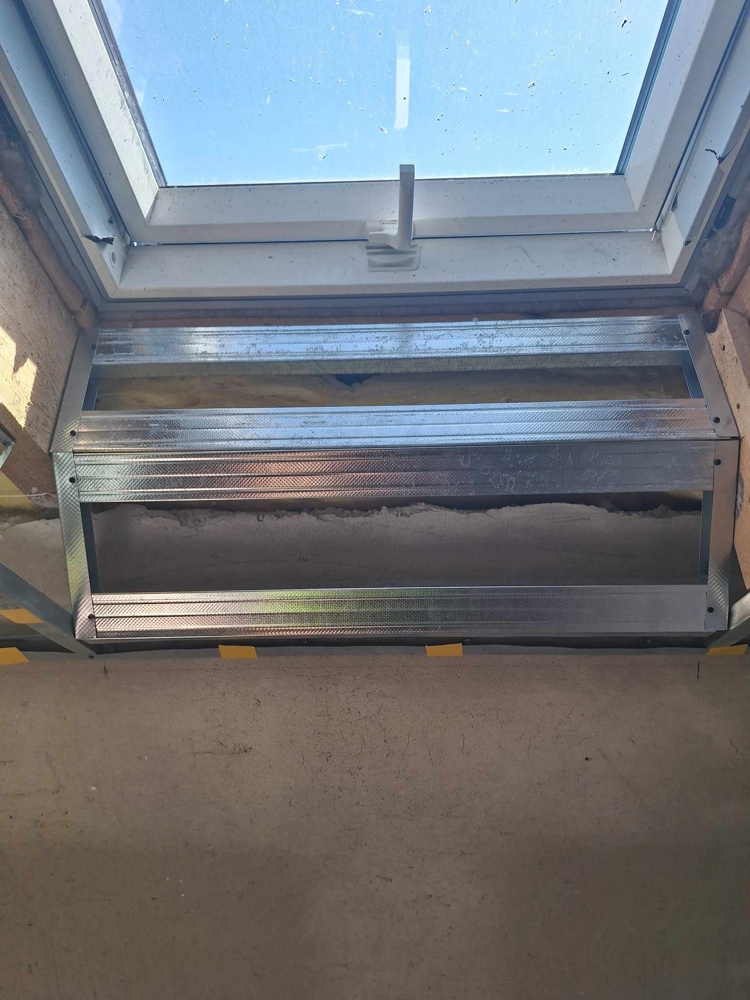 Montaż stelaża pod sufit napinany wokół okna dachowego, widoczne profile metalowe i częściowo wypełniona przestrzeń izolacją. Ujęcie z dołu, w tle błękitne niebo.