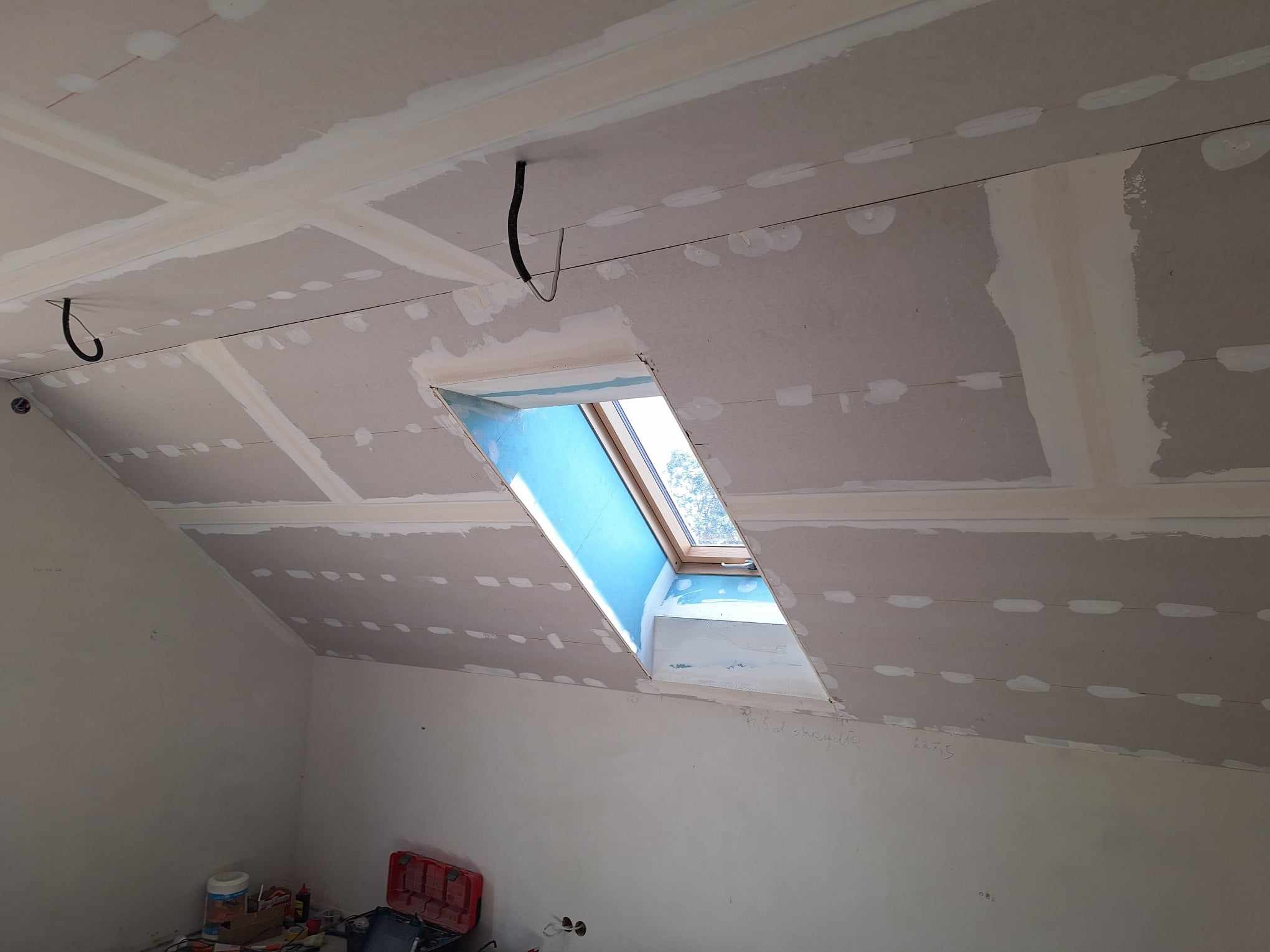 Poddasze w trakcie wykończenia: płyty gipsowo-kartonowe na skosach, okno dachowe z niebieską folią ochronną, widoczne przewody, narzędzia w tle.