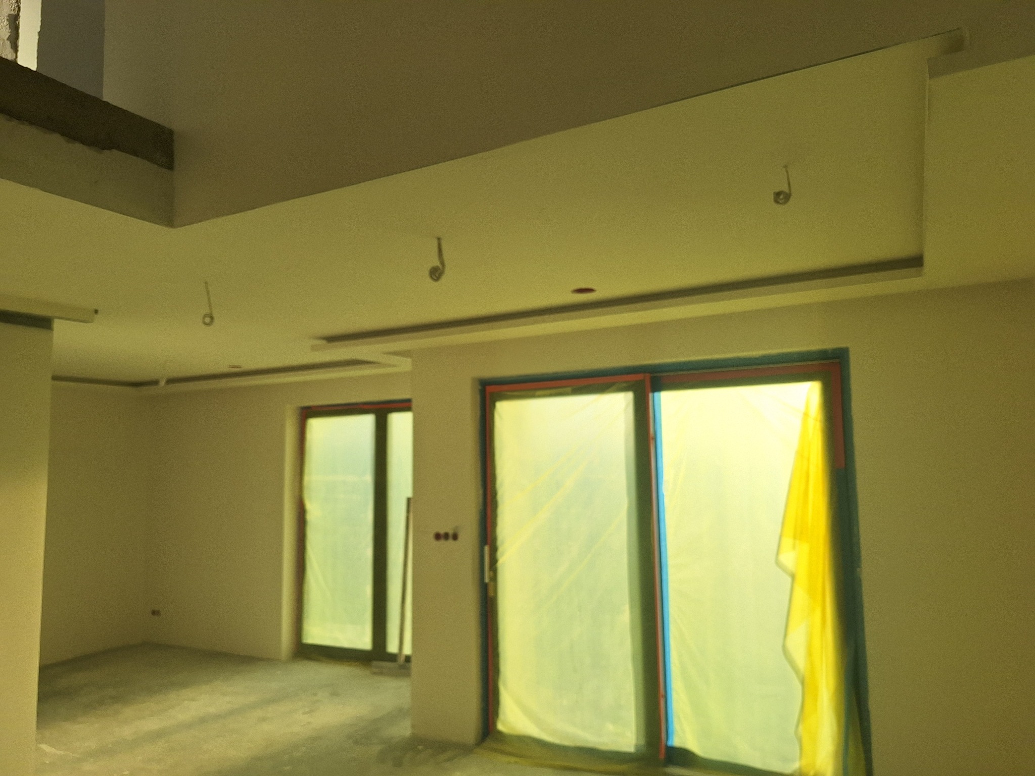 Wnętrze w trakcie wykończenia: sufit z podwieszanymi elementami, okna zabezpieczone folią. Widoczne kable oświetleniowe i surowa podłoga. Ściany przygotowane do malowania.