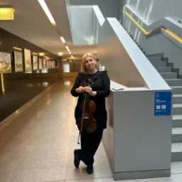 Blondwłosa kobieta w czarnej sukience trzyma skrzypce w korytarzu z obrazami i schodami.