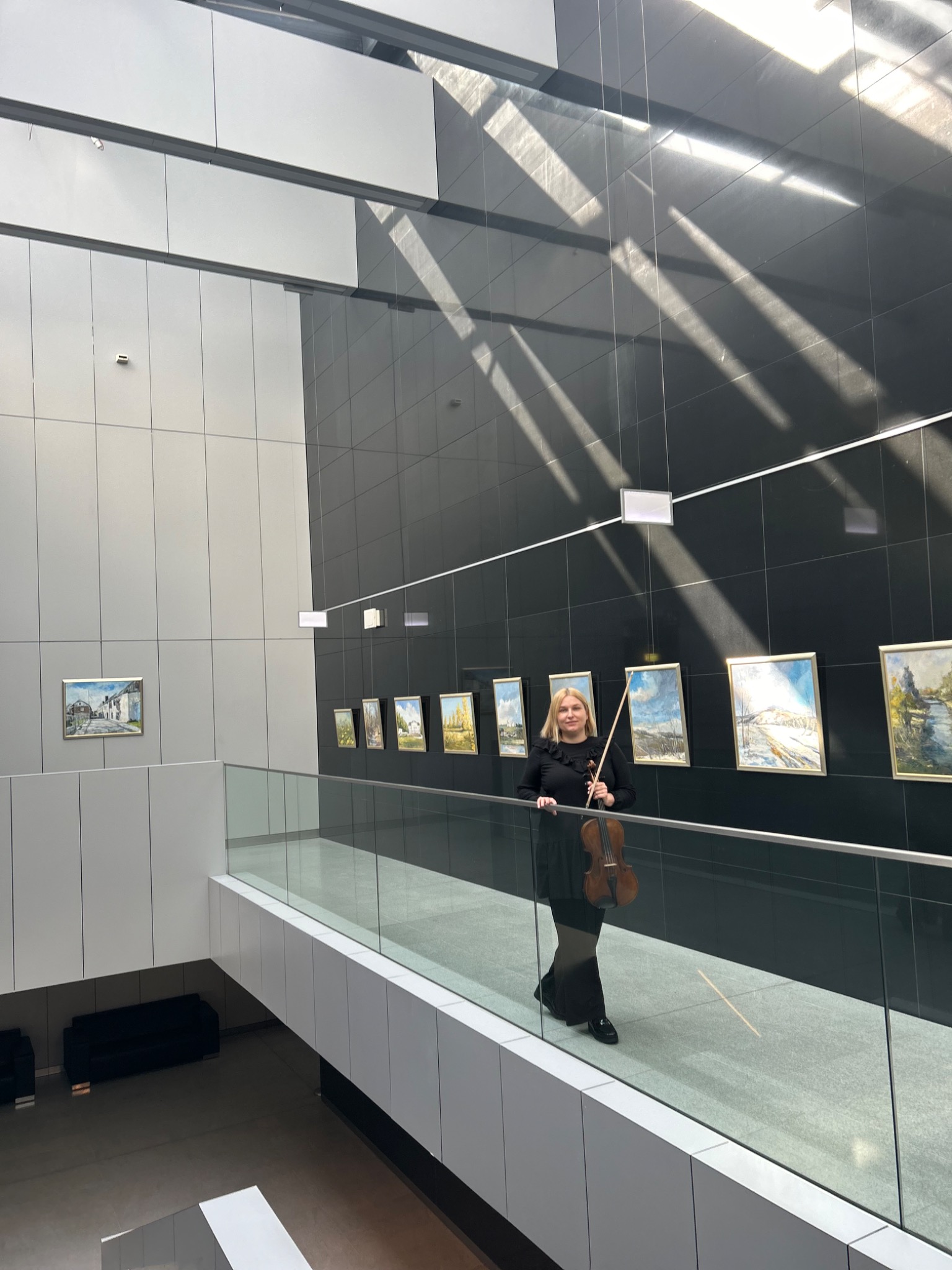 Kobieta z blond włosami, ubrana na czarno, stoi na przeszklonym balkonie z skrzypcami w dłoni, na tle ściany z czarnych płytek, na której wiszą obrazy, a przez okno wpadają promienie słoneczne.